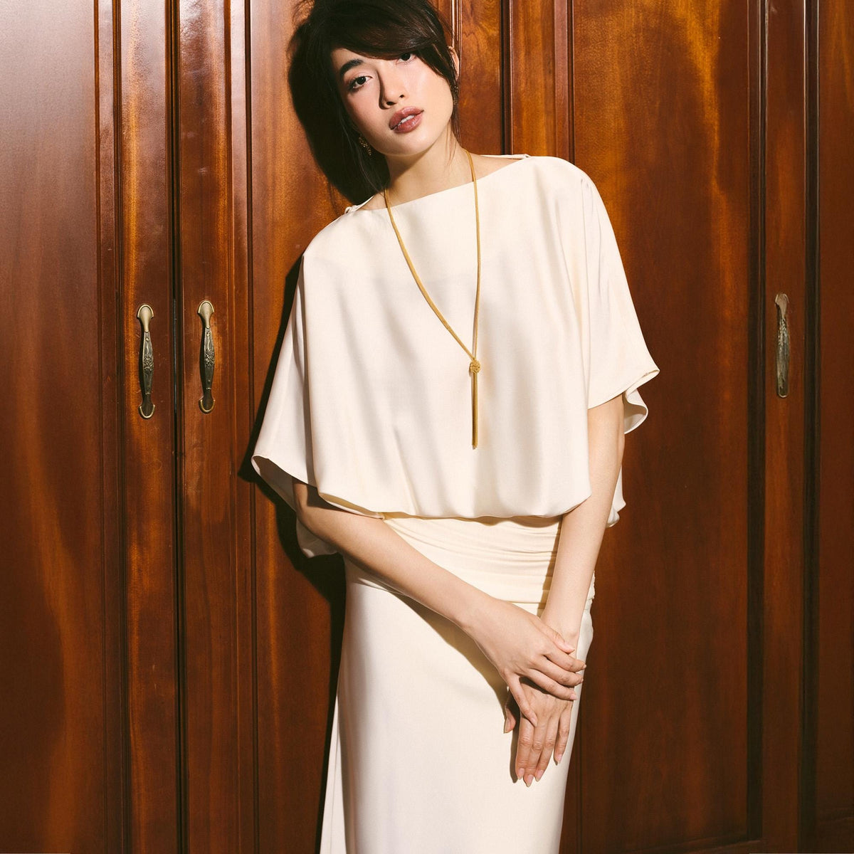 May include: A woman wearing a cream-colored dress with a matching top and skirt. The top has short sleeves and a draped design. A long gold necklace hangs down the front. The background is a dark wood door.