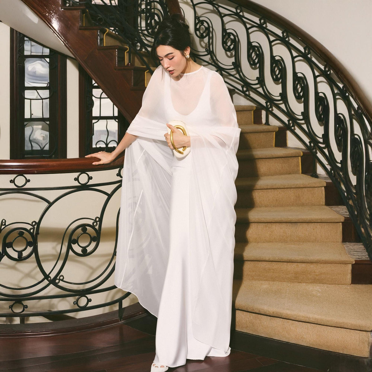 May include: A woman in a flowing white dress and sheer shawl descends a curved staircase with a dark wood banister and ornate black metal railing. She holds a white clutch with a gold handle. The stairs are carpeted in a neutral color.