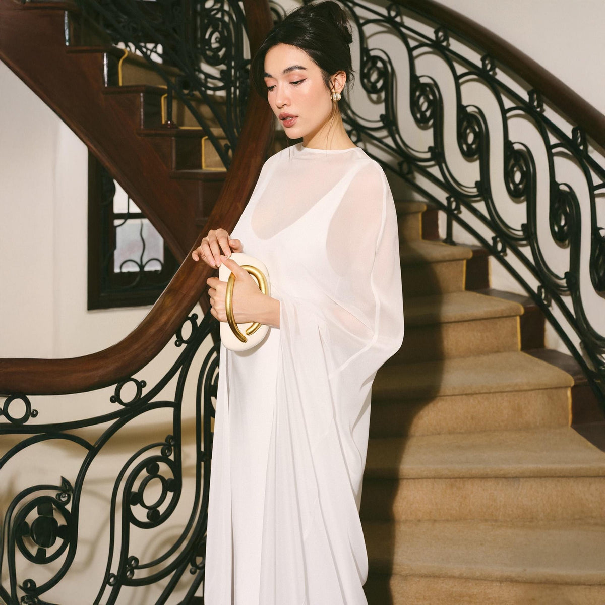 May include: A white, flowing dress with sheer sleeves and a matching clutch with a gold handle. The dress is worn on a staircase with a dark wooden banister and wrought iron railing.