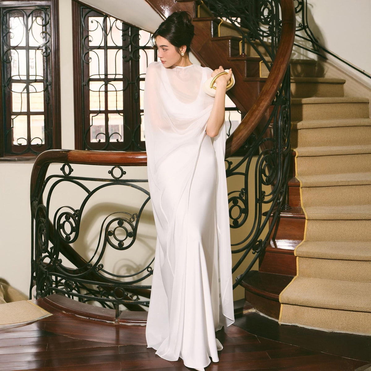 May include: A white, floor-length gown with a flowing cape. The dress has a fitted bodice and a flared skirt. The cape is sheer and drapes over the shoulders. A gold clutch is held in the hand. The background includes a staircase and decorative railing.