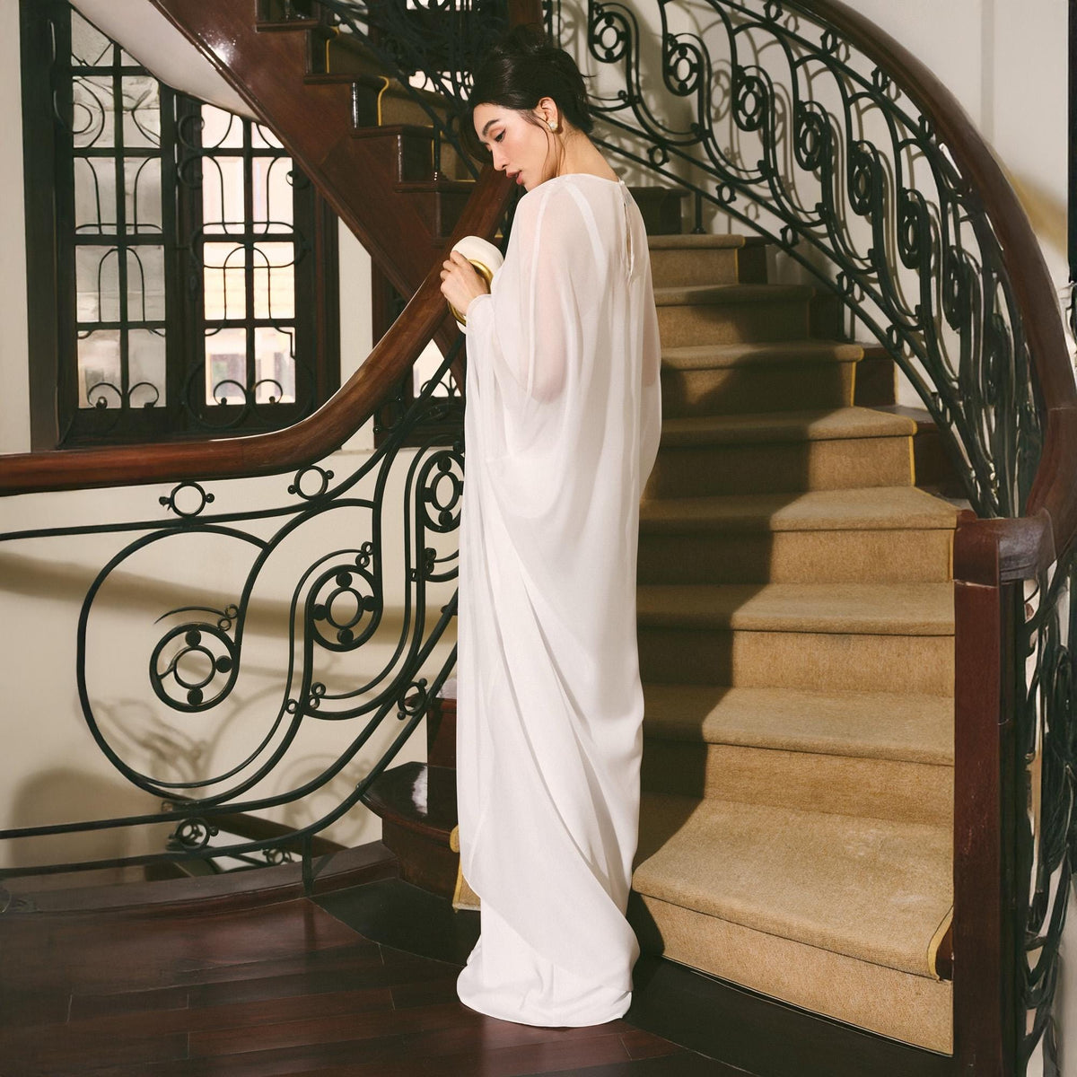 May include: A flowing, floor-length white dress with wide sleeves. The dress is made of a lightweight, sheer fabric. The woman is standing on a staircase with a dark wood banister and ornate wrought iron railing. The stairs are covered in a beige carpet.