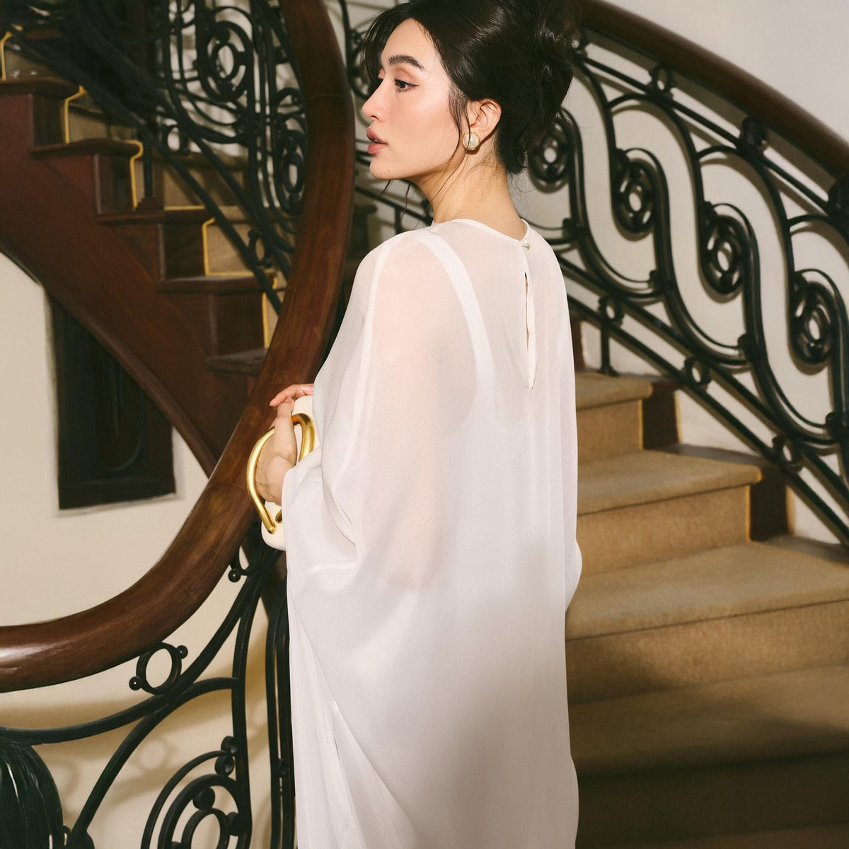 May include: A sheer white dress with a flowing silhouette. The dress has a simple neckline and long sleeves. The background features a wooden staircase with a dark brown handrail and black metal detailing.