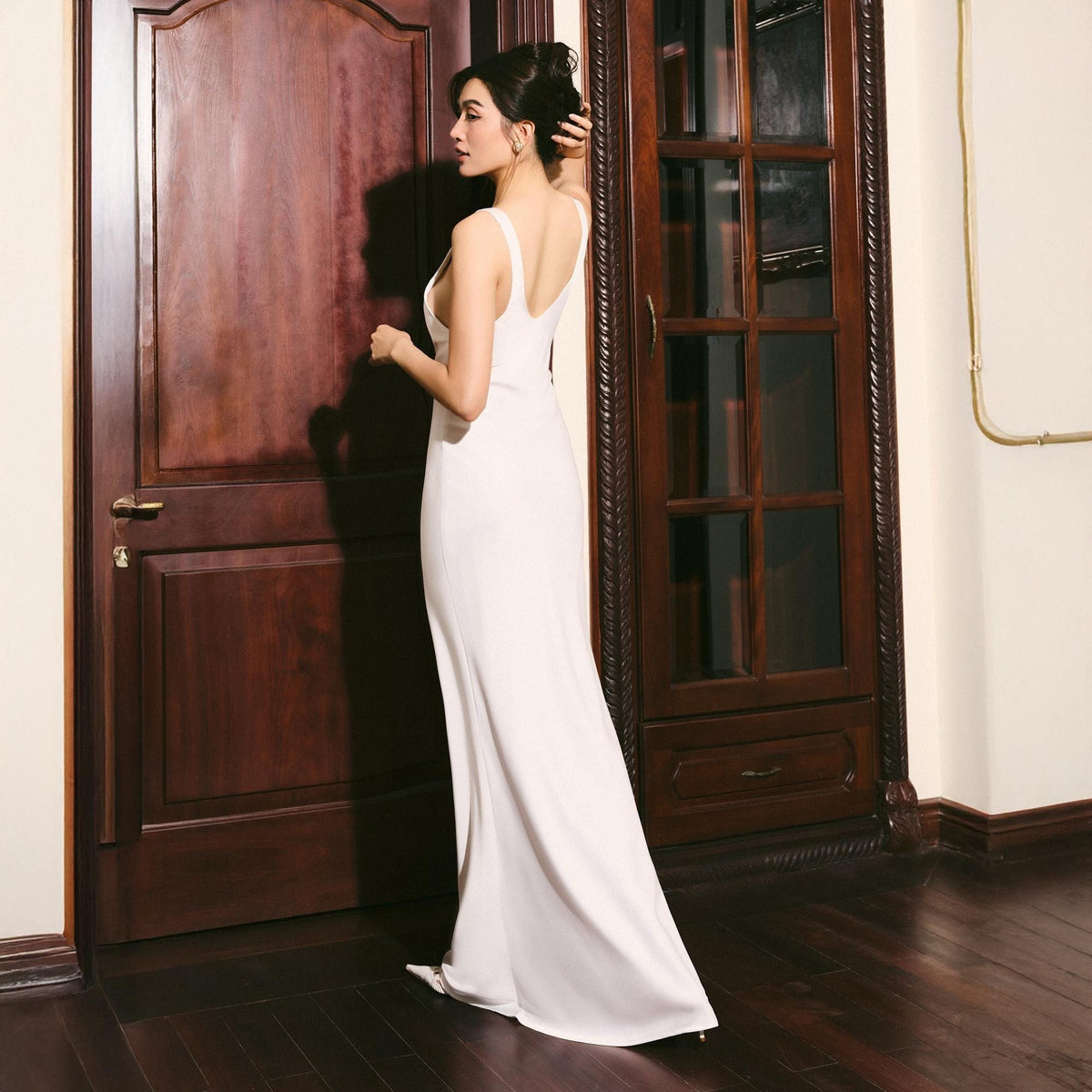 May include: A woman in a white, floor-length gown with a low back and spaghetti straps stands near a dark wooden door. The dress has a simple, elegant design. The woman's hair is styled up, and she is looking to the side.