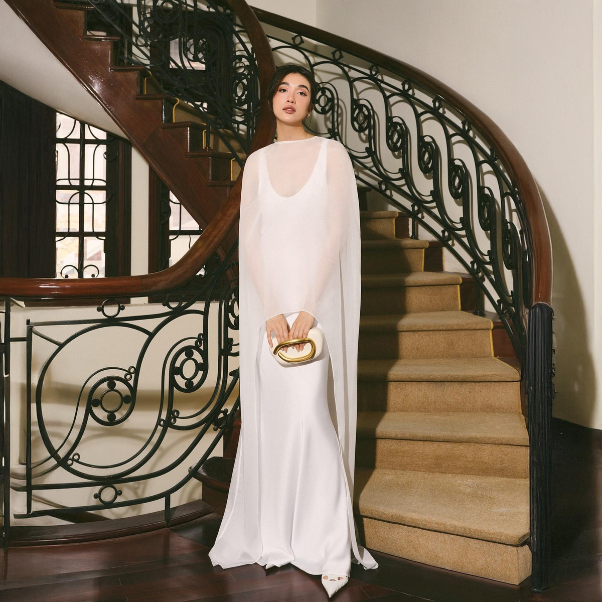 May include: A woman in a white, floor-length gown with a sheer cape stands on a staircase. She holds a small, white clutch with a gold handle. The staircase has a dark wood banister with ornate black metalwork.