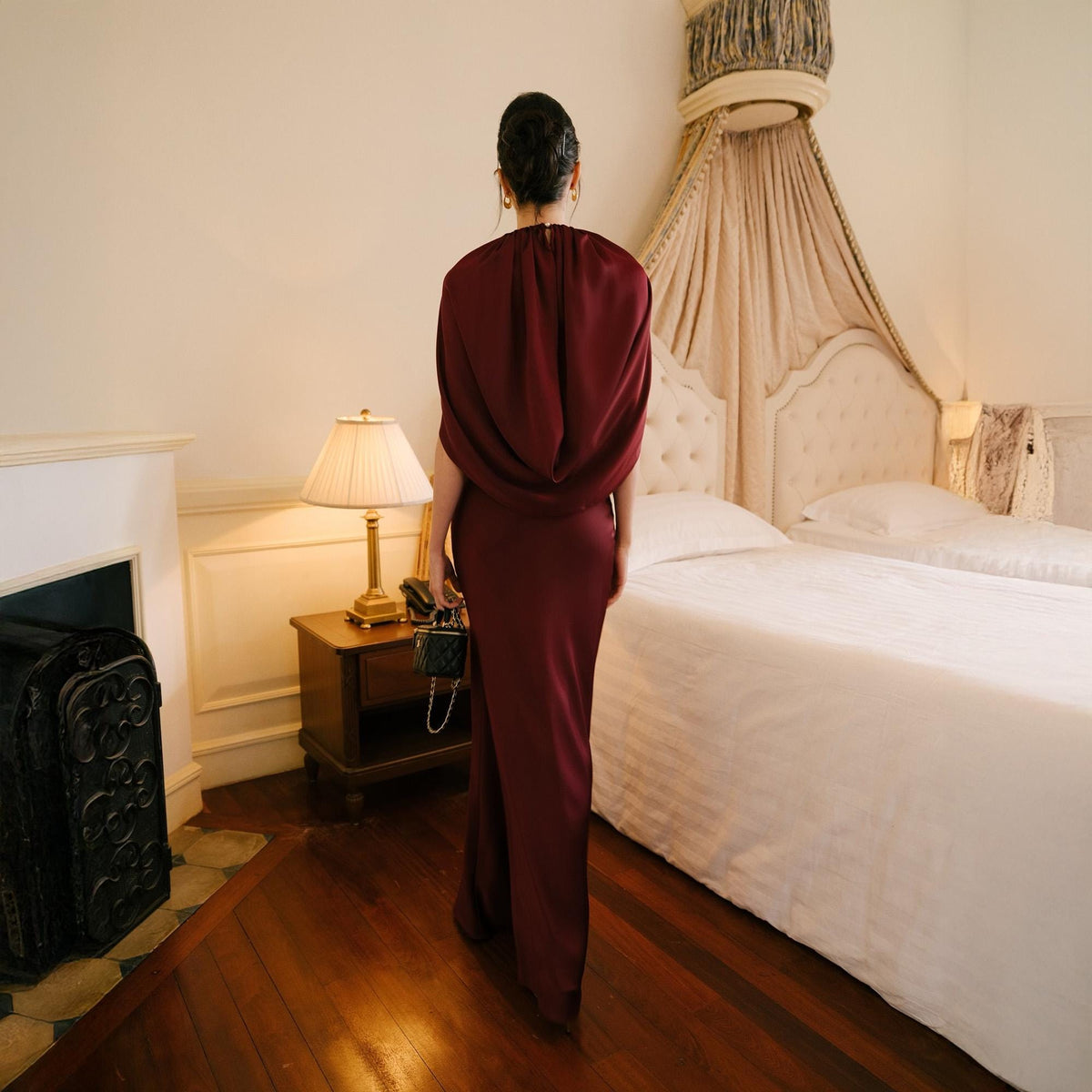 May include: A burgundy-colored, floor-length gown with a draped cowl neckline. The dress has a fitted silhouette and is worn by a person standing in a room with a bed and a fireplace. A small black handbag with a chain strap is held.