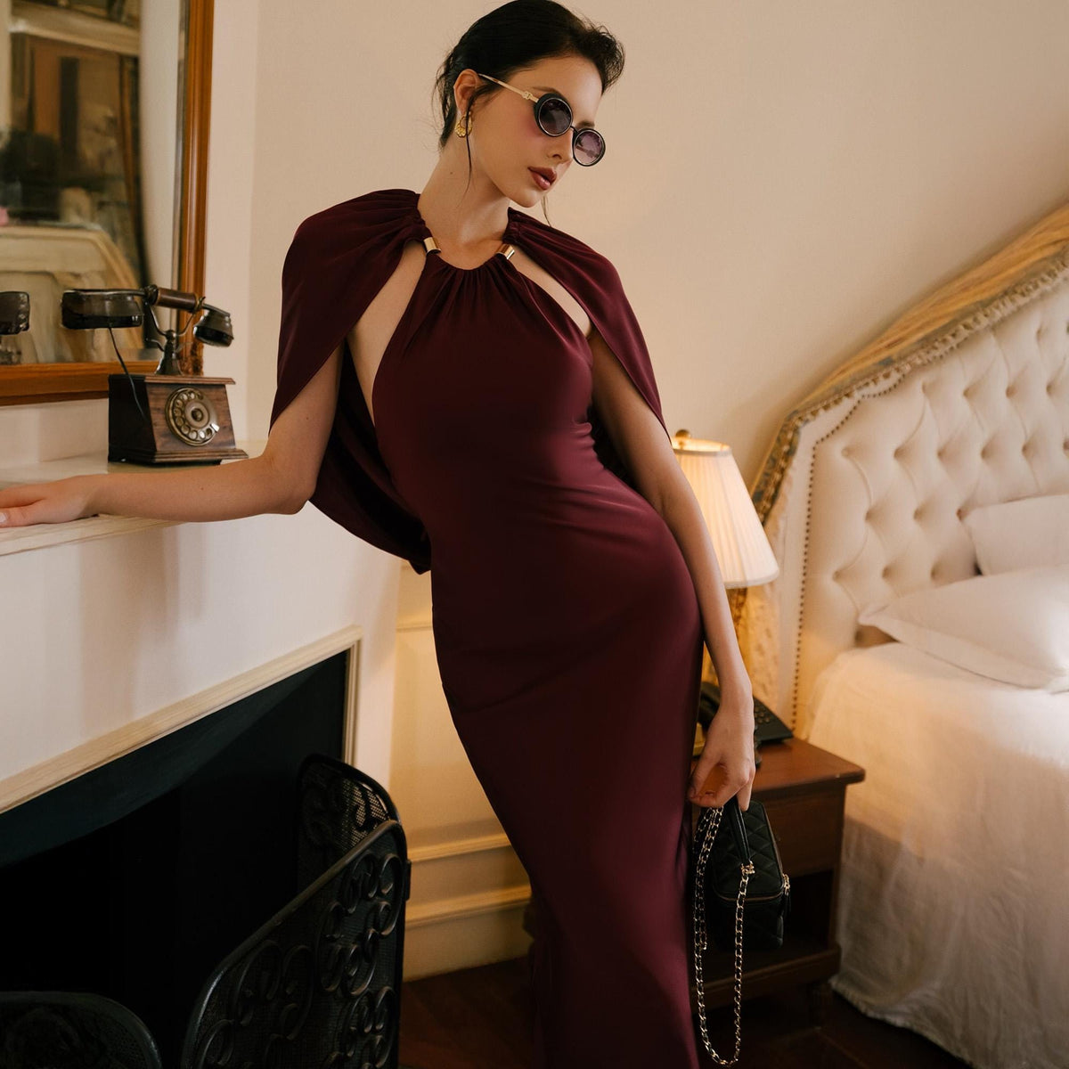 May include: A burgundy-colored formal dress with a cape-like overlay. The dress features a halter neckline with a gold-colored ring detail. The cape drapes over the shoulders. A small black purse with a chain strap is held in the hand.