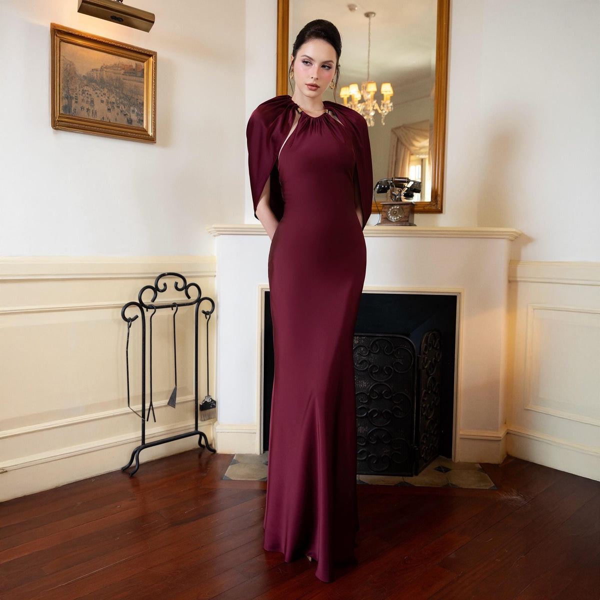 May include: A burgundy formal gown with a matching cape. The floor-length dress has a fitted silhouette that flares out slightly at the bottom. The cape is draped over the shoulders and is attached at the neckline. The background includes a fireplace and a mirror.