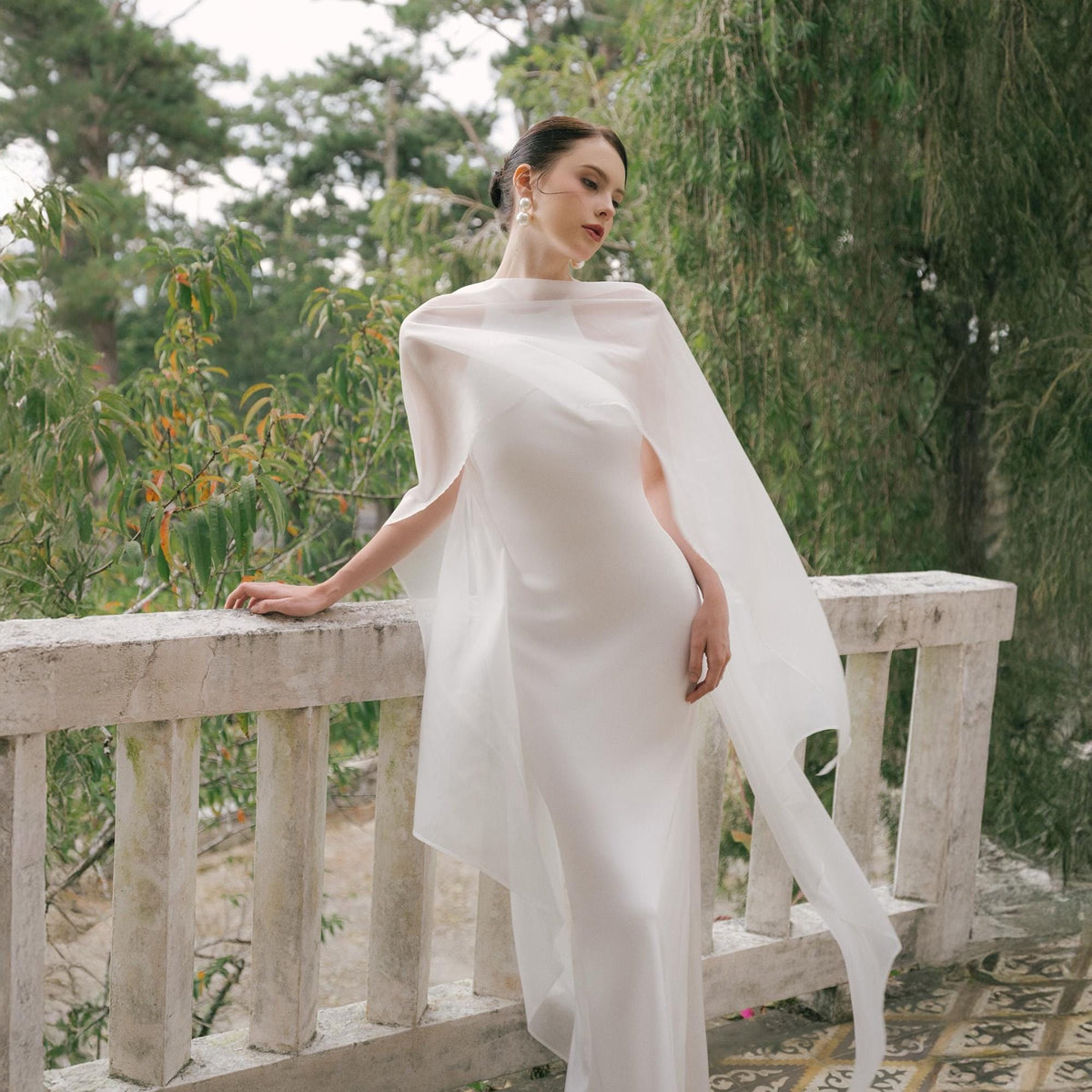 May include: A white, floor-length gown with a matching sheer cape. The dress has a simple, elegant design, and the cape drapes over the shoulders. The background features greenery and a white railing.