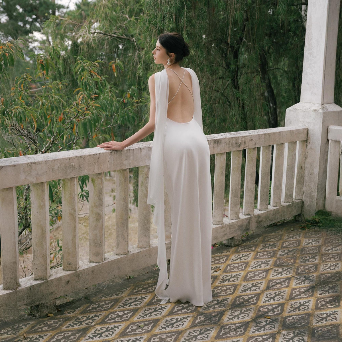 May include: A white, floor-length gown with a low back and flowing cape-like sleeves. The dress features a simple, elegant design, perfect for a formal event. The model is standing on a balcony overlooking a lush green landscape.