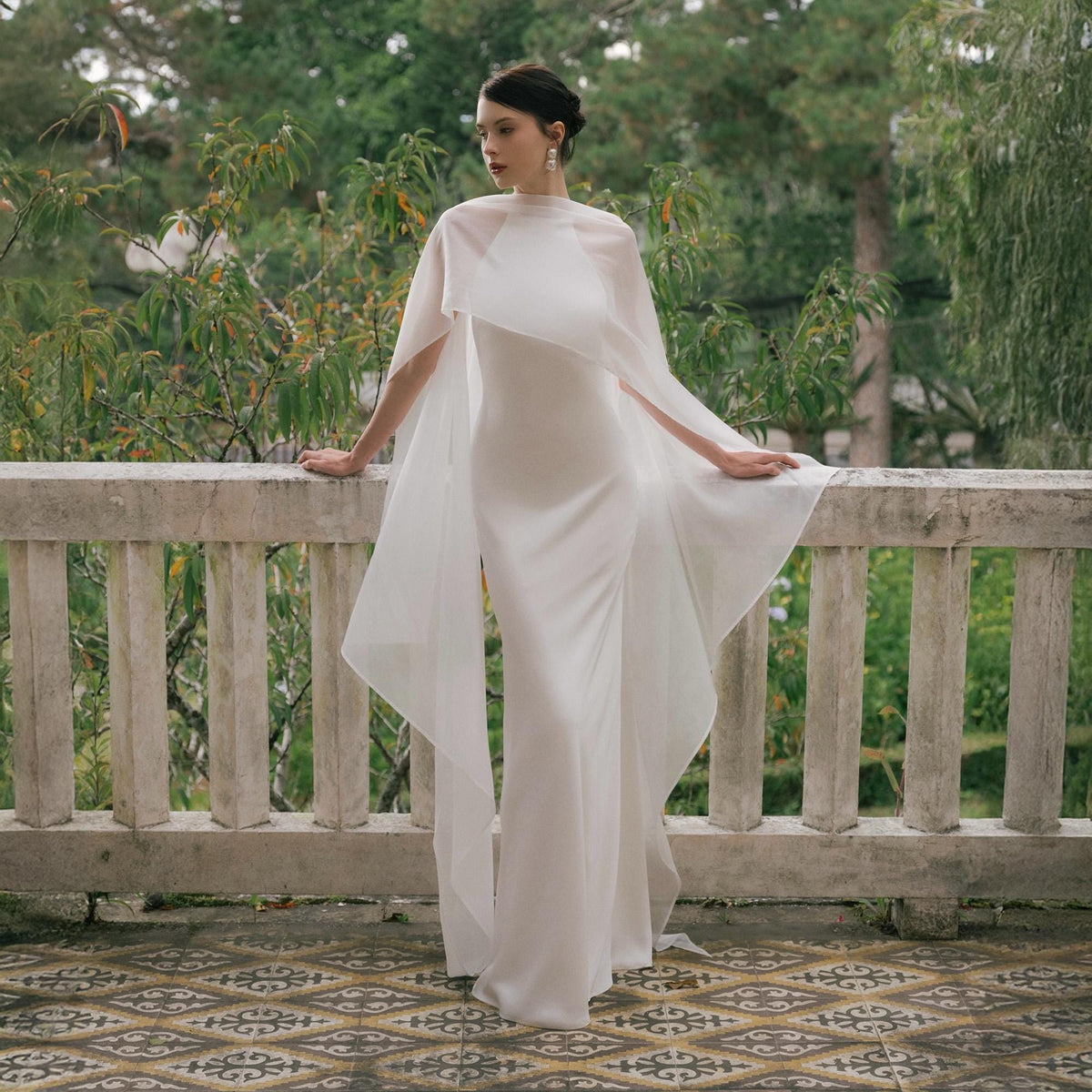 May include: A white, floor-length gown with a high neckline and a flowing cape. The cape drapes over the shoulders and arms, creating a dramatic silhouette. The dress is simple and elegant, perfect for a special occasion.