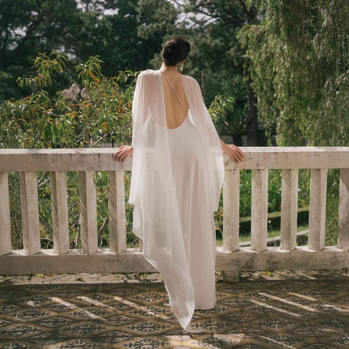 May include: A white wedding dress with a flowing cape, standing on a balcony overlooking a lush green landscape. The dress has a low back and delicate straps. The cape drapes elegantly, creating a romantic and ethereal look. The balcony has a white railing.