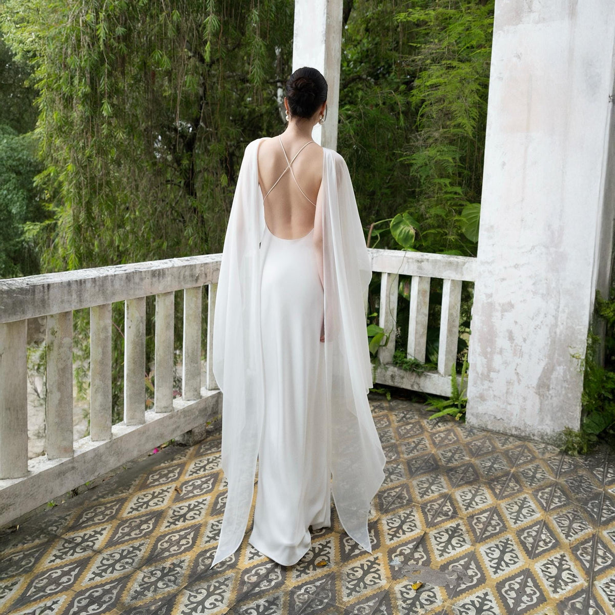 May include: A white wedding dress with a low back and flowing cape sleeves. The dress is floor-length and the model is standing on a balcony overlooking lush greenery. The dress has a simple, elegant design.