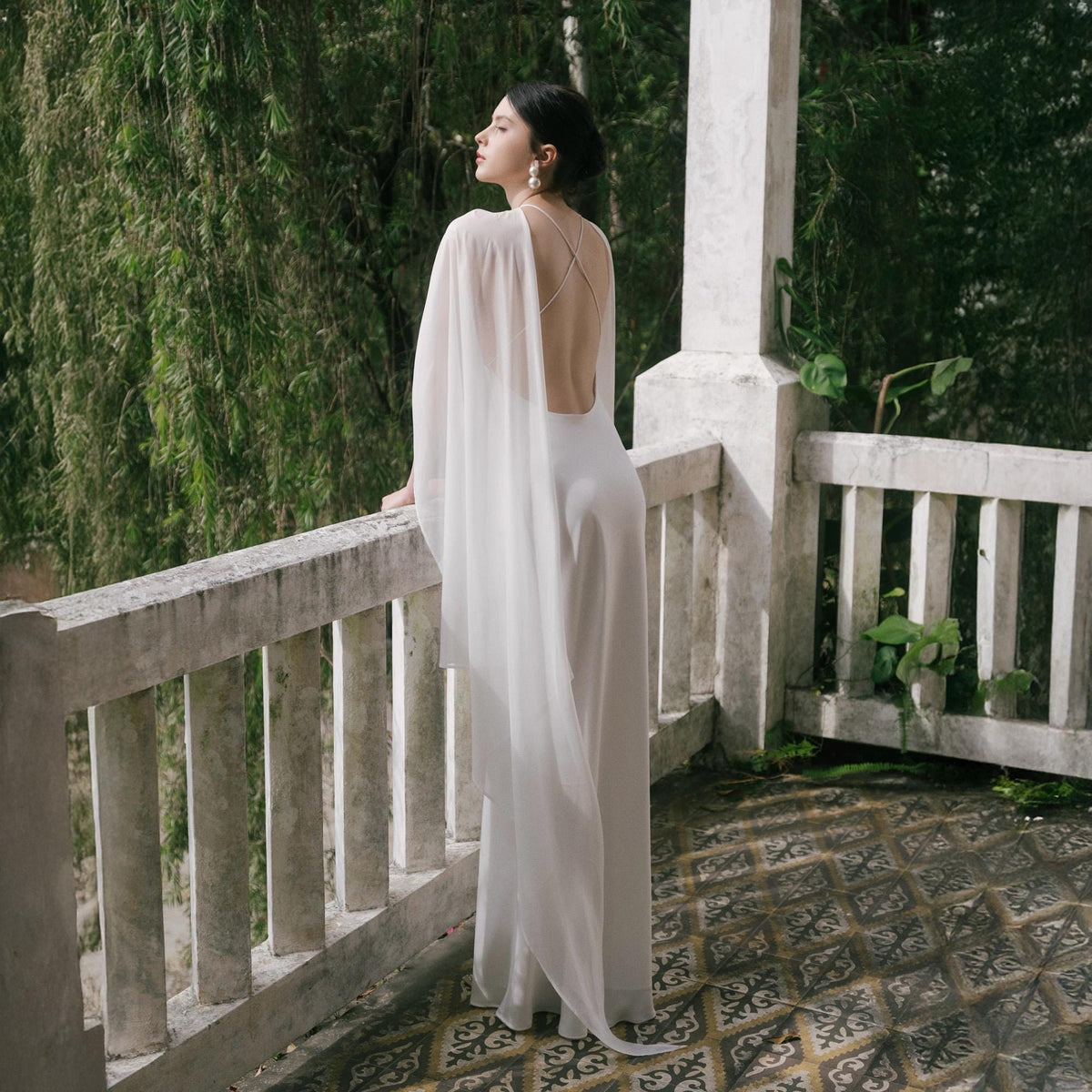 May include: A white, flowing gown with a low back and sheer cape. The dress is worn on a balcony with a stone railing, overlooking lush greenery. The dress has a simple, elegant design.