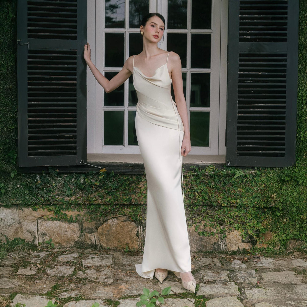 May include: A cream-colored, floor-length gown with a cowl neckline and spaghetti straps. The dress has a fitted silhouette and a slight train. The model is standing in front of a window with black shutters and a stone wall covered in greenery.
