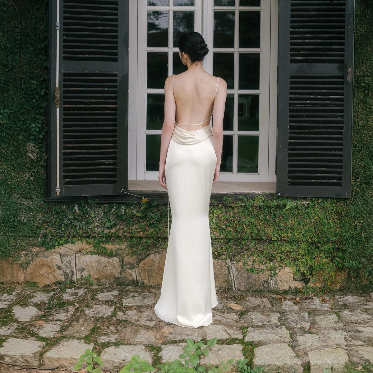 May include: A backless ivory-colored gown with a flowing mermaid silhouette. The dress features delicate spaghetti straps and a cowl neckline. The model is standing in front of a window with black shutters, surrounded by greenery.
