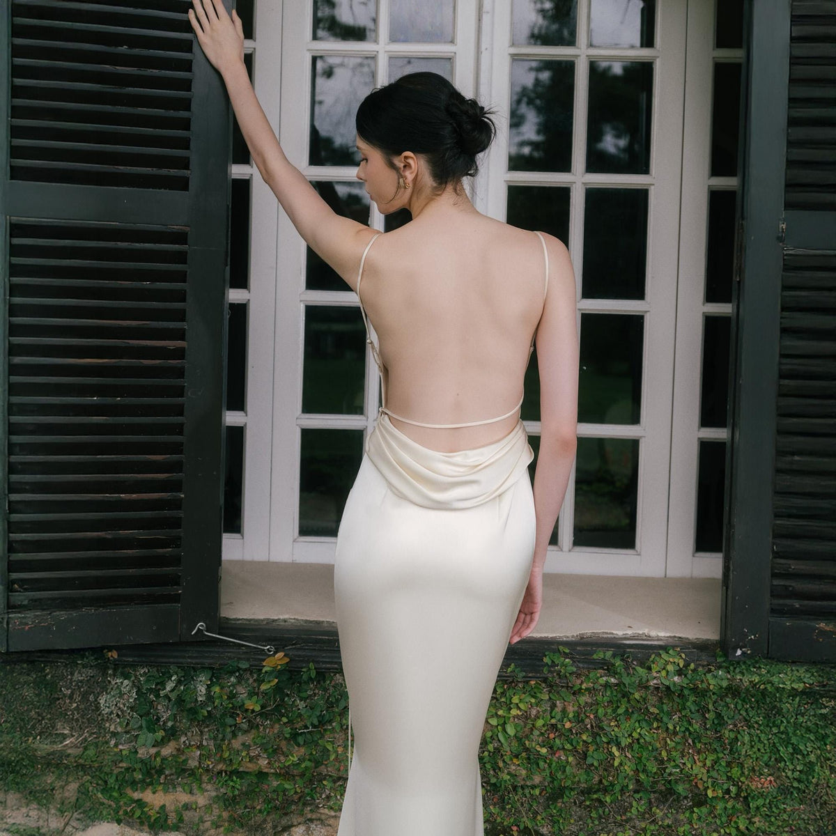 May include: A cream-colored, backless, floor-length dress with spaghetti straps. The dress has a cowl neckline and a fitted silhouette. The model is standing in front of a white door with dark green shutters.