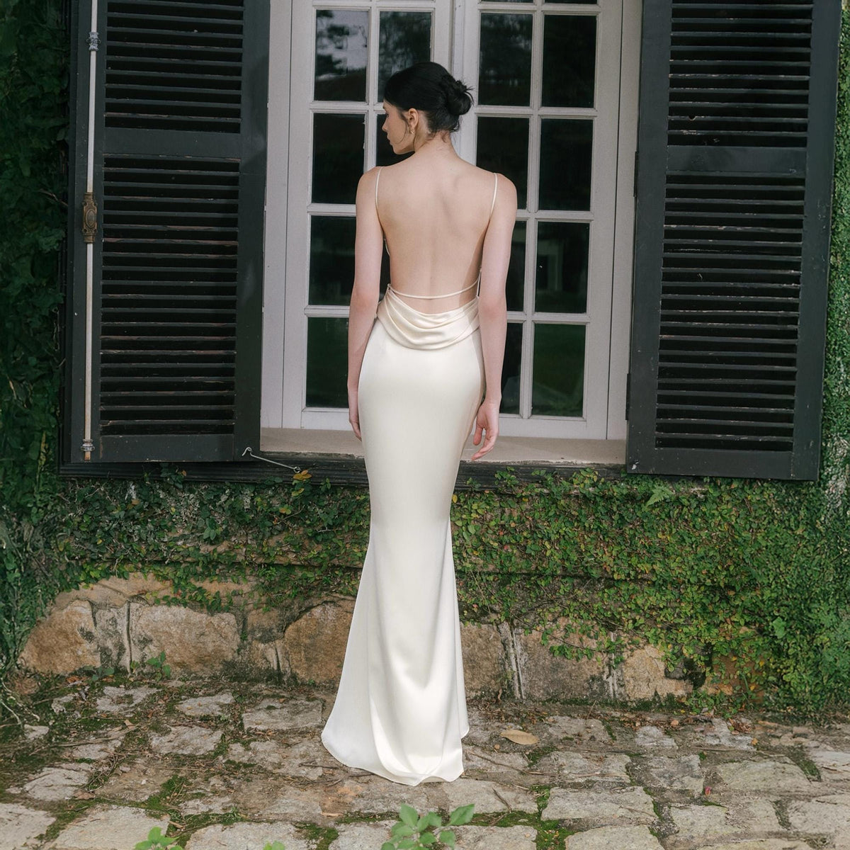 May include: A cream-colored, backless, floor-length gown with spaghetti straps. The dress features a cowl neckline and a fitted silhouette. The model is standing in front of a window with black shutters and a stone wall covered in greenery.