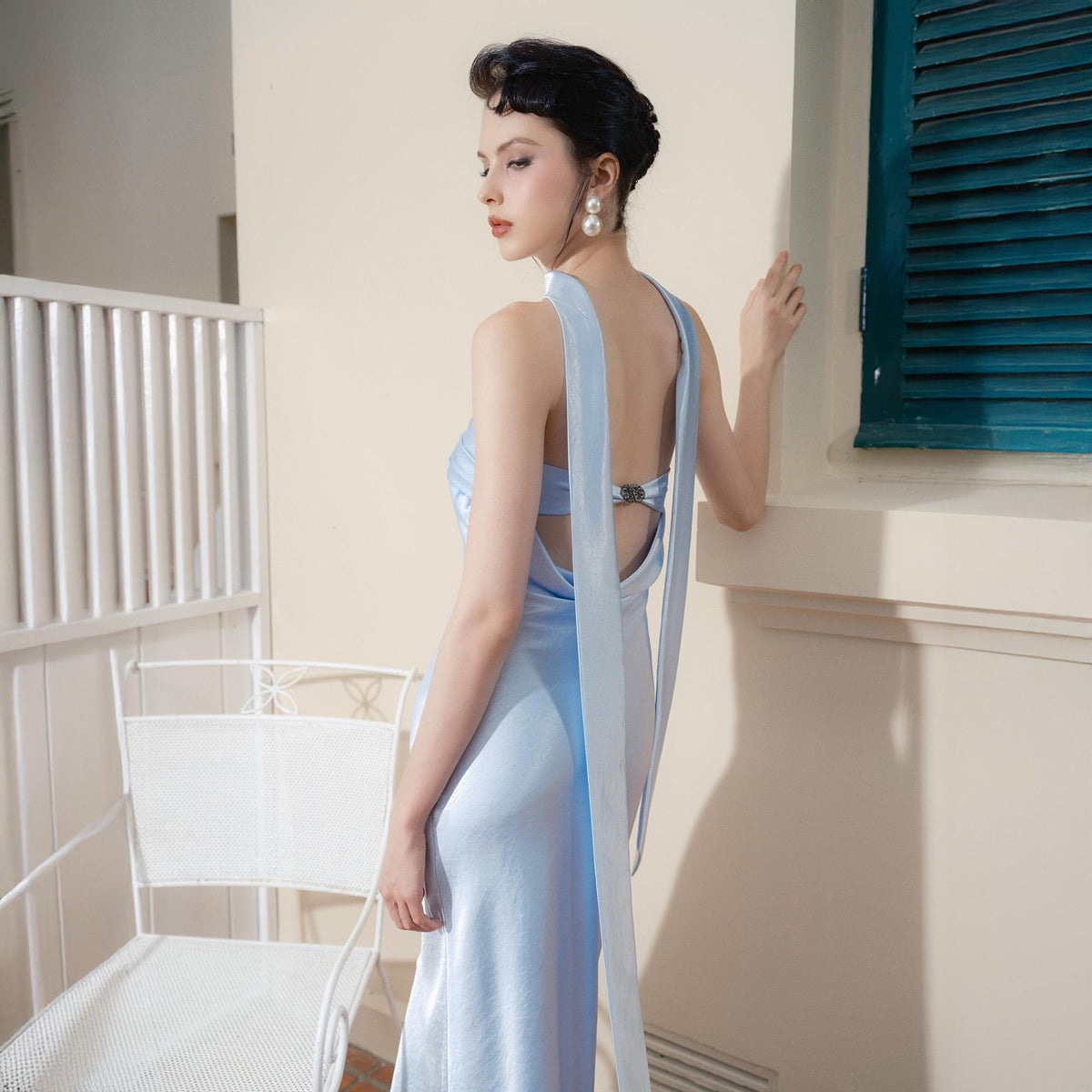 May include: A light blue satin dress with a halter neckline and open back. The dress features long, flowing fabric that drapes down the back. The model is wearing pearl earrings and has dark hair styled in an updo.