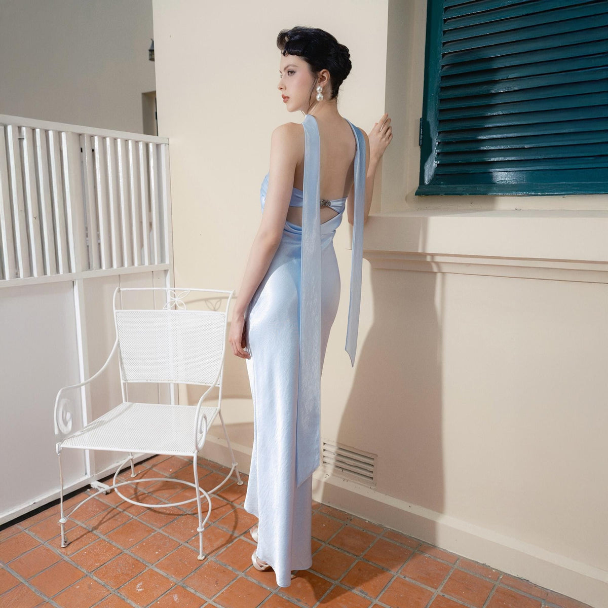 May include: A light blue, floor-length dress with a low back and long, flowing straps. The dress is made of a soft, silky material. A white metal chair is in the background. The model is wearing white sandals.