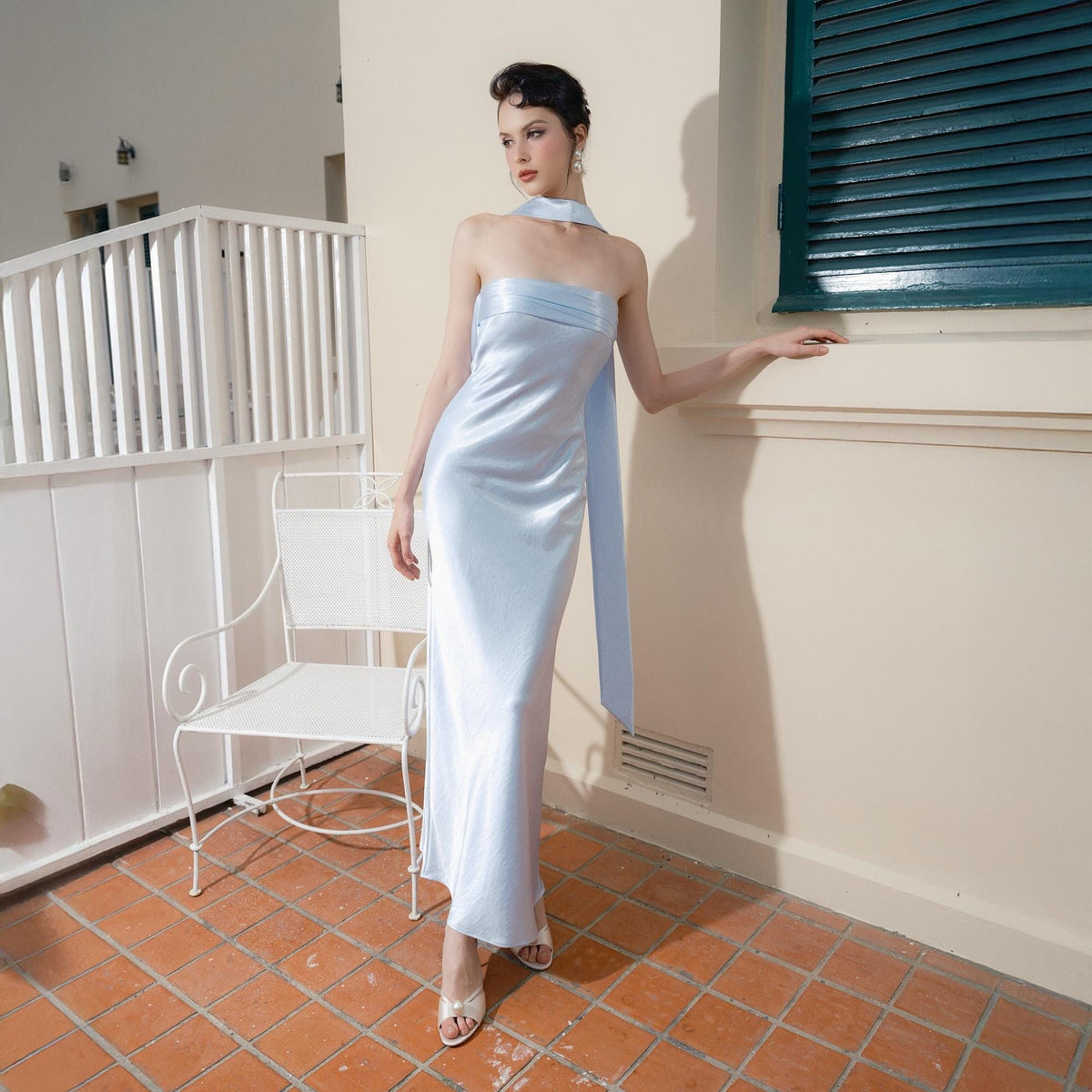 May include: A light blue satin gown with a strapless design and a flowing scarf detail. The dress is floor-length and has a subtle sheen. The model is wearing white heels. The background includes a white chair and a window with green shutters.