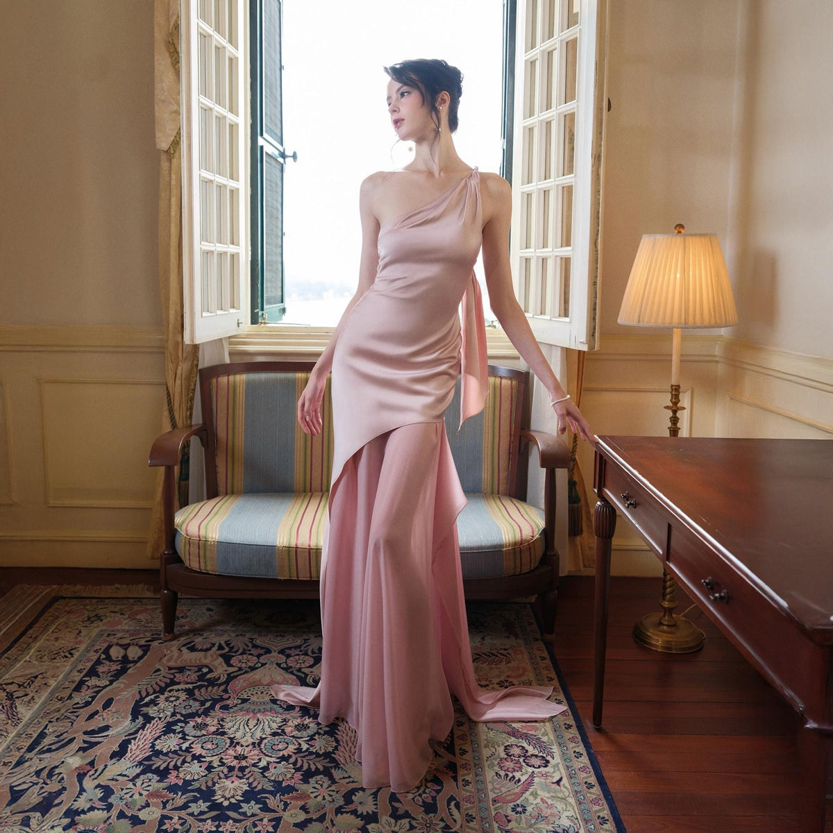 May include: A light pink, one-shoulder formal gown with a flowing skirt and a draped accent. The dress features a high slit and a long train. The gown is made of a silky material and is suitable for special occasions.