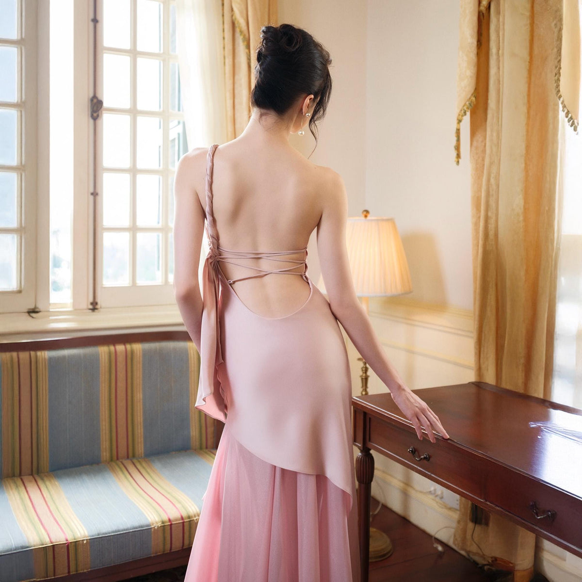 May include: A light pink, backless formal dress with a flowing skirt. The dress features a halter-style strap and crisscross back detailing. The skirt has a layered design, and the dress appears to be made of a soft, flowing fabric. The dress is suitable for formal events.