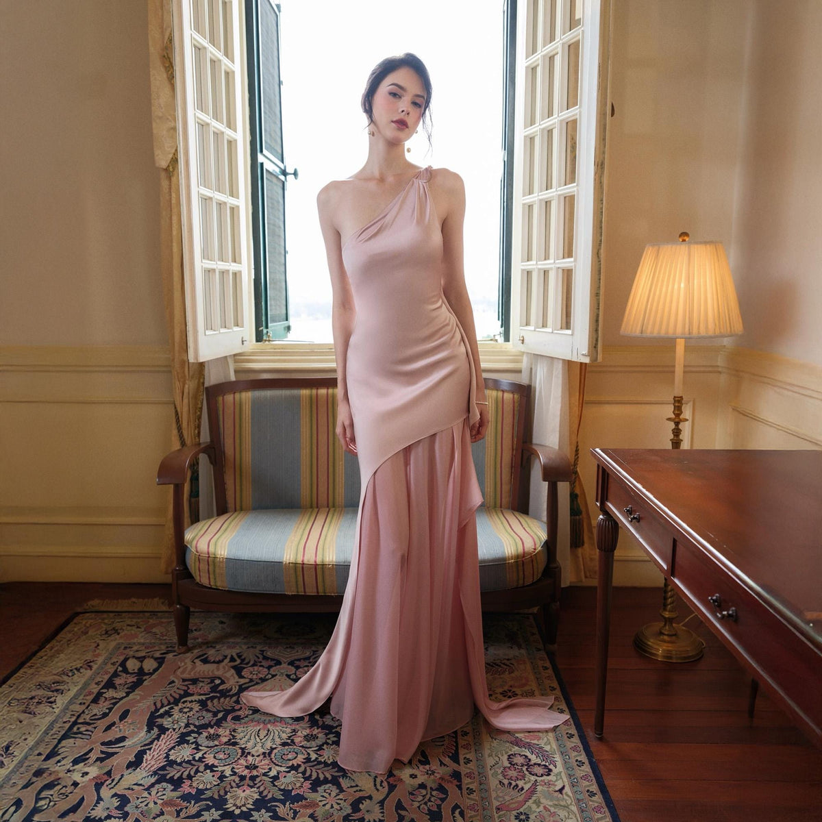 May include: A blush pink, one-shoulder formal gown with a flowing skirt. The dress features a draped bodice and a cascading asymmetrical skirt. The model is standing in front of a window, with a vintage-style sofa and a patterned rug in the background.