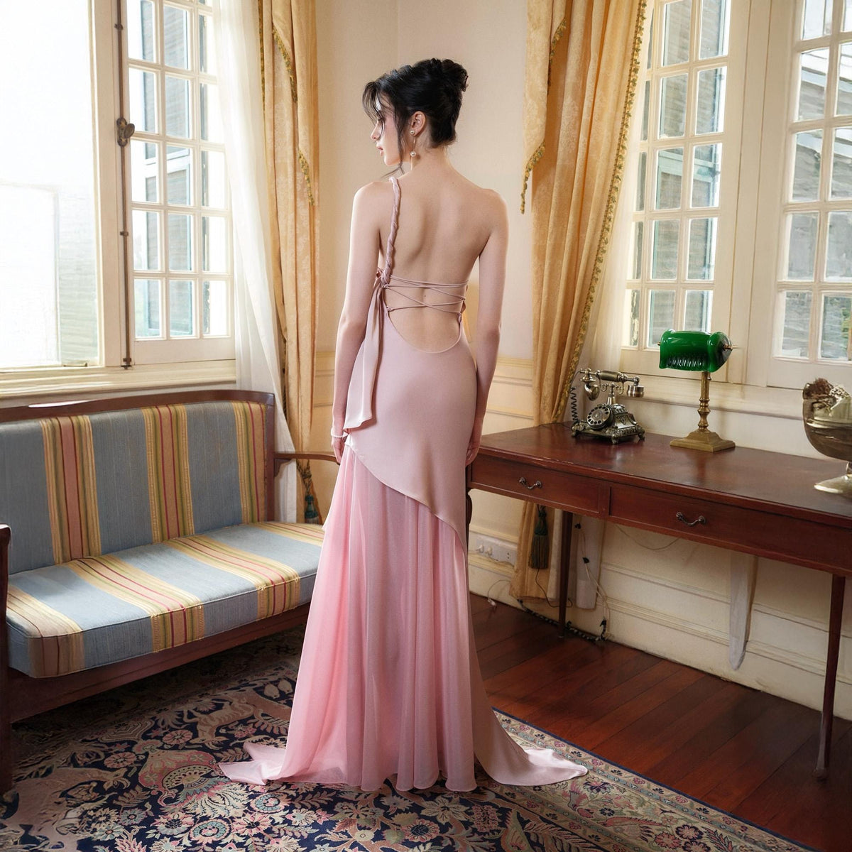May include: A light pink formal gown with a flowing skirt and open back. The dress features a one-shoulder strap and a draped overlay. The gown is floor-length and made of a soft, flowing fabric. The background includes a window, curtains, and a wooden table.