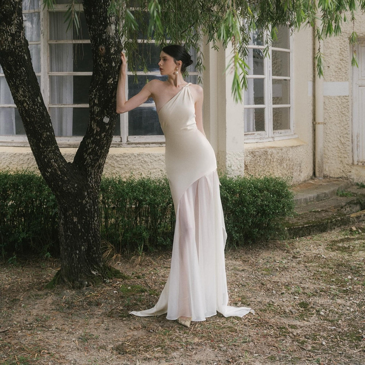 May include: An off-white, one-shoulder gown with a flowing, sheer skirt. The dress has a fitted bodice and a draped design. The model is standing near a tree in an outdoor setting. The dress is suitable for a wedding or formal event.