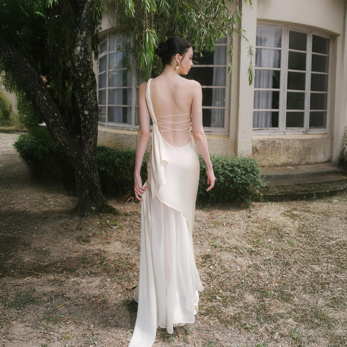 May include: A cream-colored, one-shoulder gown with a flowing skirt and open back. The dress features delicate spaghetti straps across the back and a draped fabric detail. The model is standing outdoors near a building with large windows.