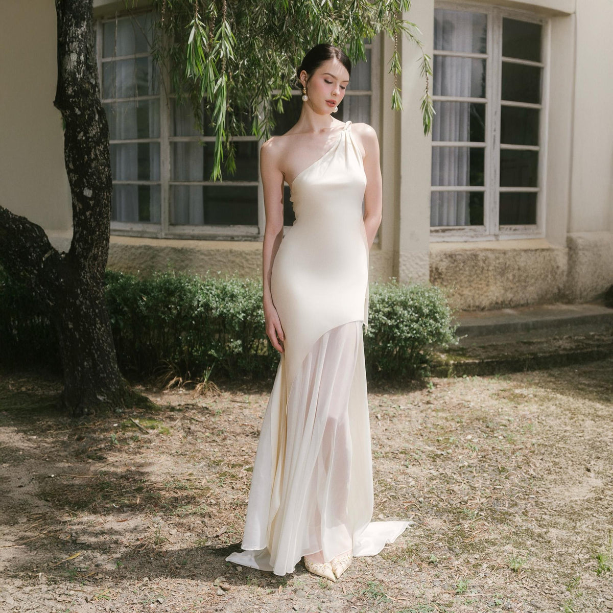 May include: A cream-colored, one-shoulder gown with a flowing skirt. The dress features a fitted bodice and a sheer, asymmetrical overlay. The model is wearing gold-colored shoes. The dress is suitable for a formal event.