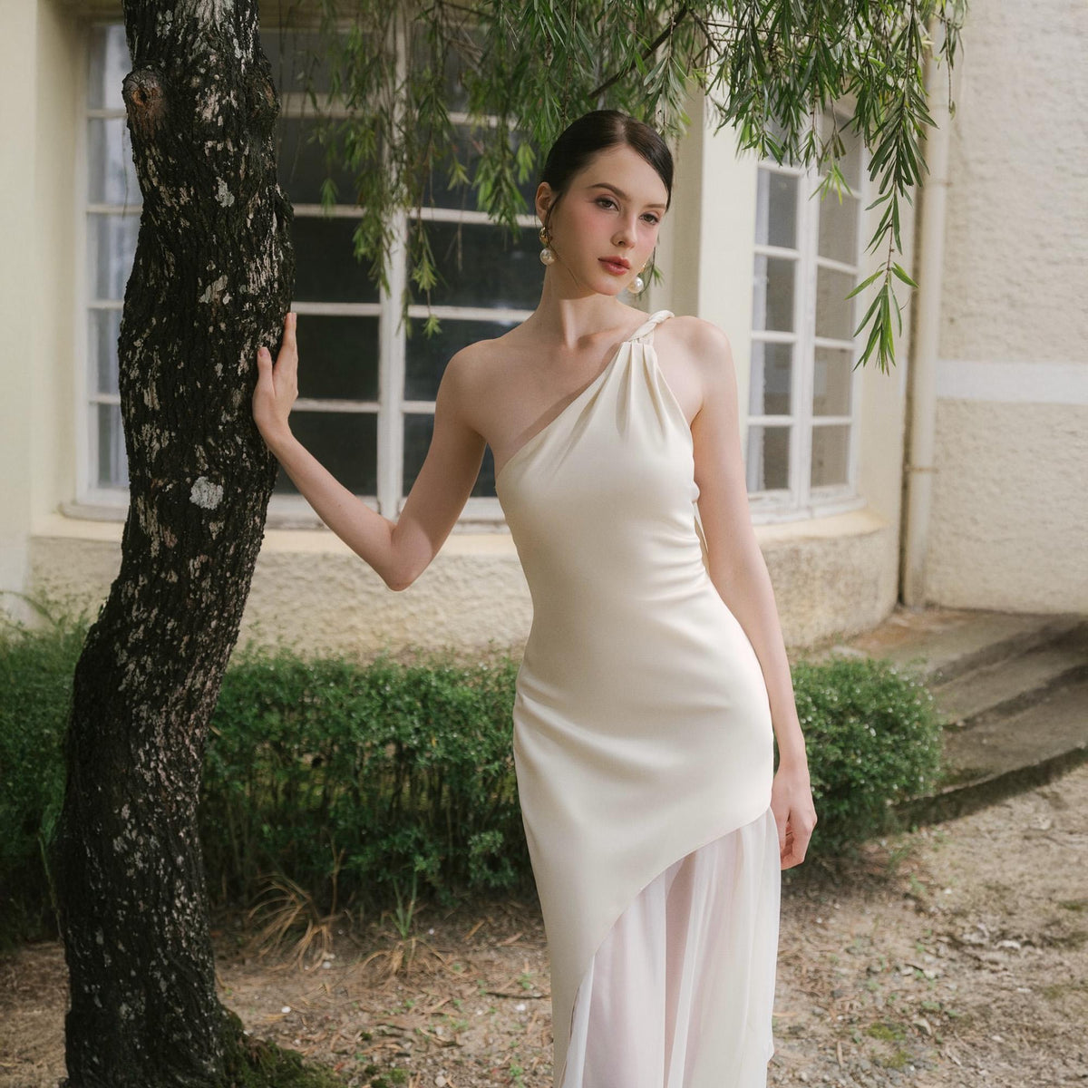 May include: Ivory one-shoulder dress with a draped neckline and asymmetrical hem. The dress features a flowing, sheer overlay on the skirt. The model is standing near a tree in an outdoor setting, with a building and greenery in the background.