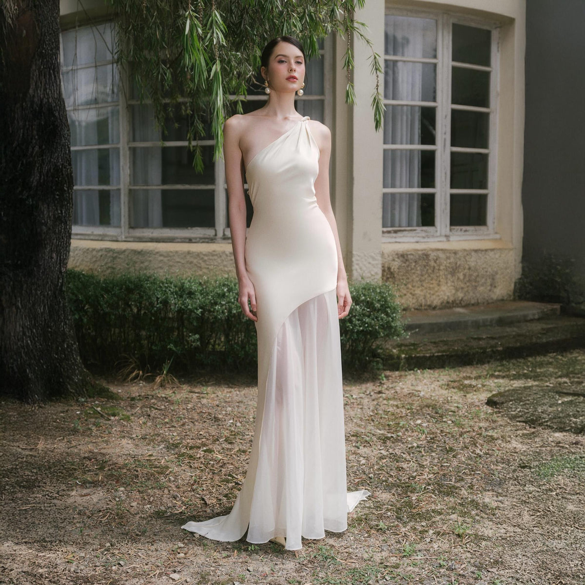 May include: An ivory-colored, one-shoulder gown with a flowing skirt. The dress features a fitted bodice and a sheer, asymmetrical overlay on the skirt. The model is standing outdoors in front of a building with large windows.