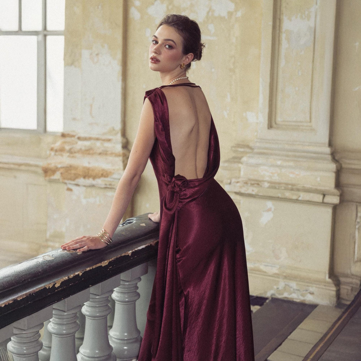 May include: A burgundy satin gown with a low back and draped neckline. The dress features a flowing skirt and is accessorized with a delicate bracelet. The model is leaning on a dark railing in a vintage setting.