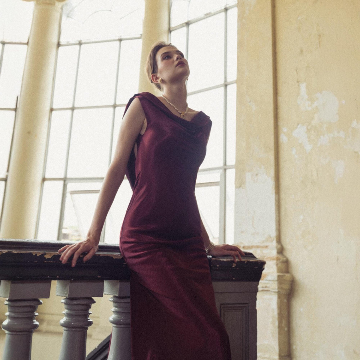 May include: A burgundy-colored, sleeveless dress with a cowl neckline. The dress is worn by a person leaning on a dark wooden railing. A pearl necklace and gold bracelet are visible. The background features a large window and aged walls.