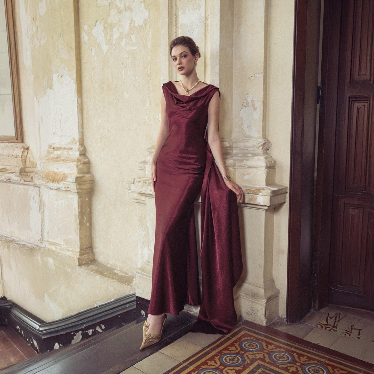 May include: A burgundy-colored, floor-length gown with a cowl neckline and a flowing, draped panel. The dress is made of a shiny fabric and is worn with gold-colored heels. The setting is an interior with architectural details.