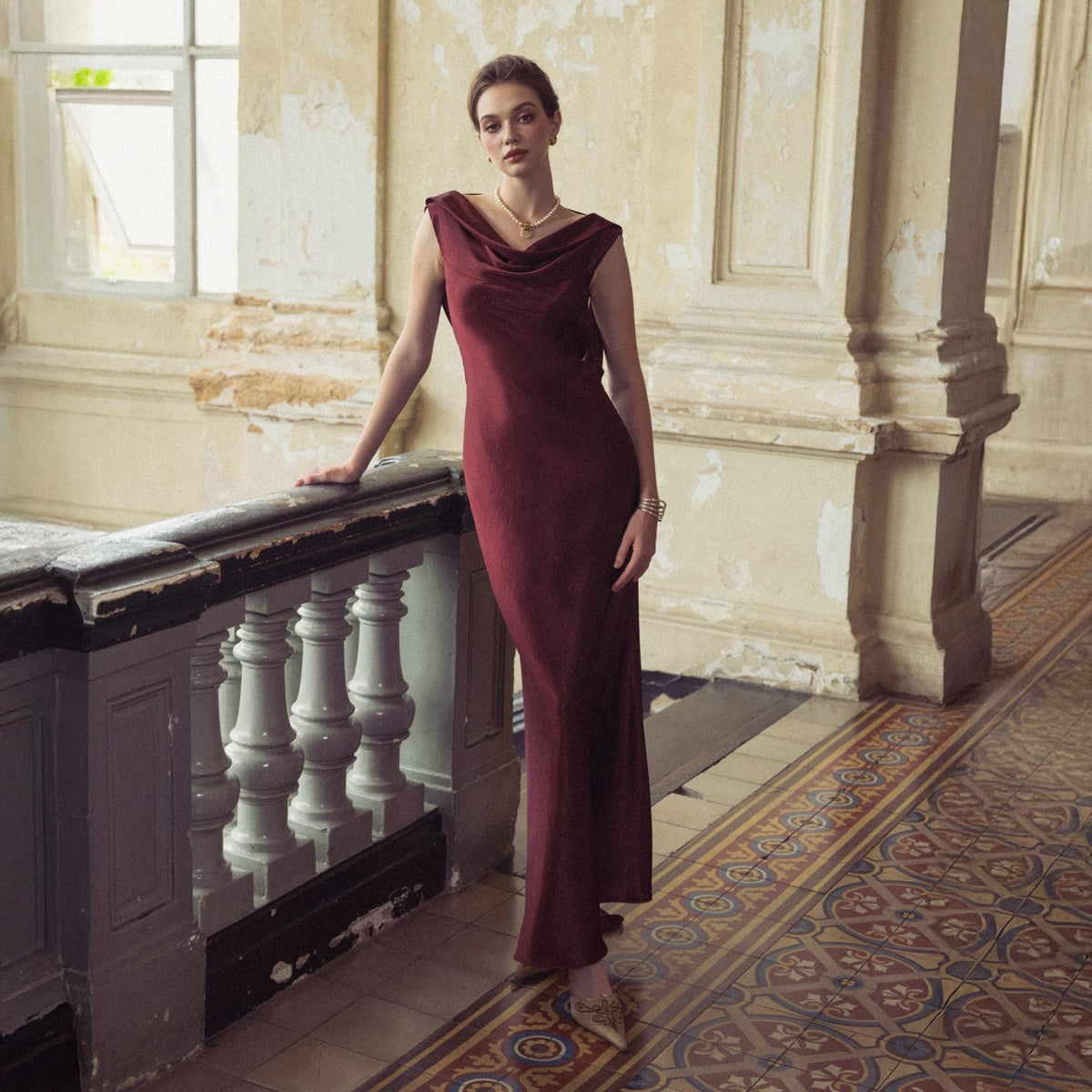 May include: A burgundy-colored, floor-length dress with a cowl neckline. The dress is sleeveless and has a flowing silhouette. The model is wearing a gold necklace and bracelet. The background features a vintage interior with a patterned tile floor.
