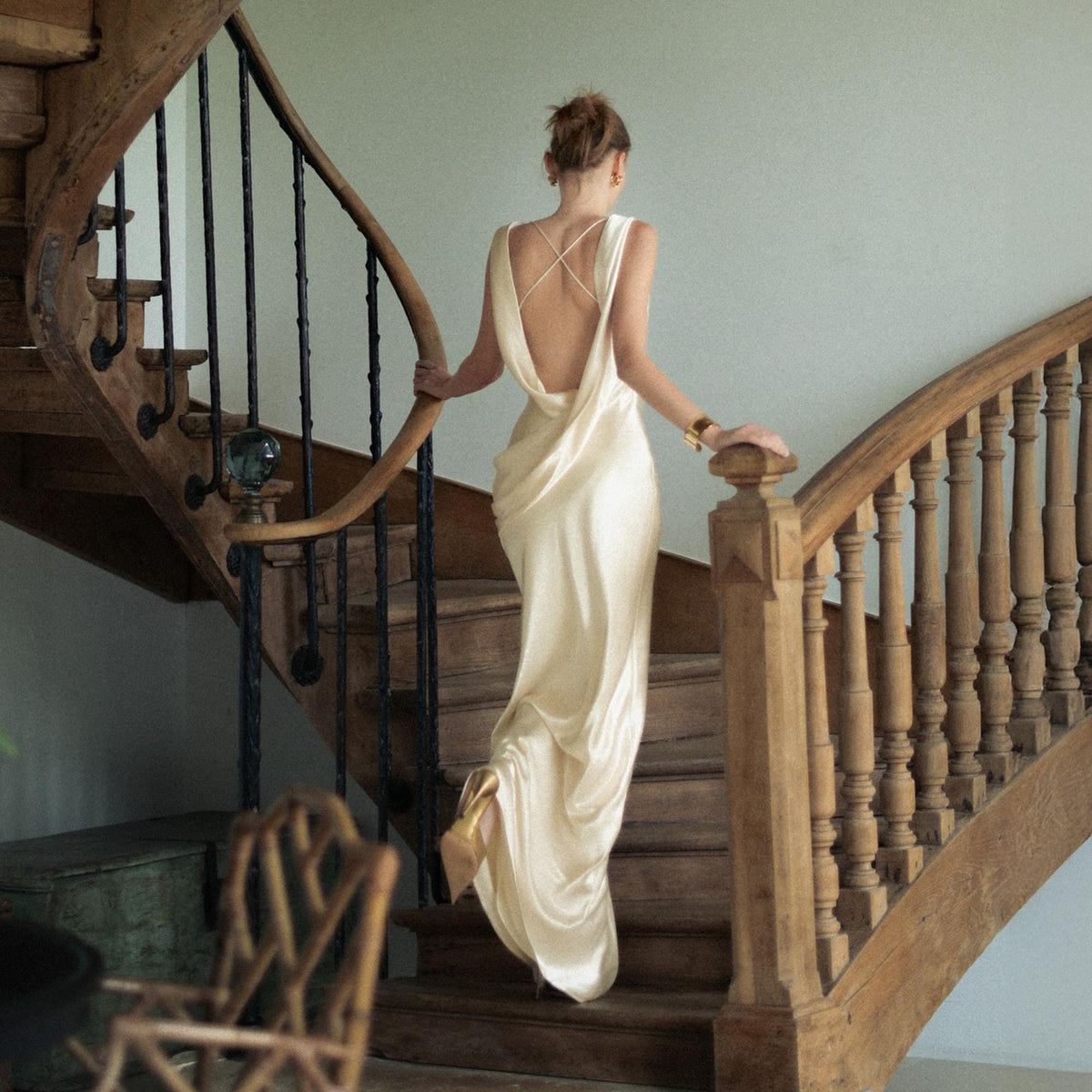 May include: A floor-length cream-colored satin gown featuring a deep cowl back, thin crisscross straps, and a draped silhouette. The fabric has a smooth, reflective finish that pools slightly at the hem, creating a fluid and elegant appearance.
