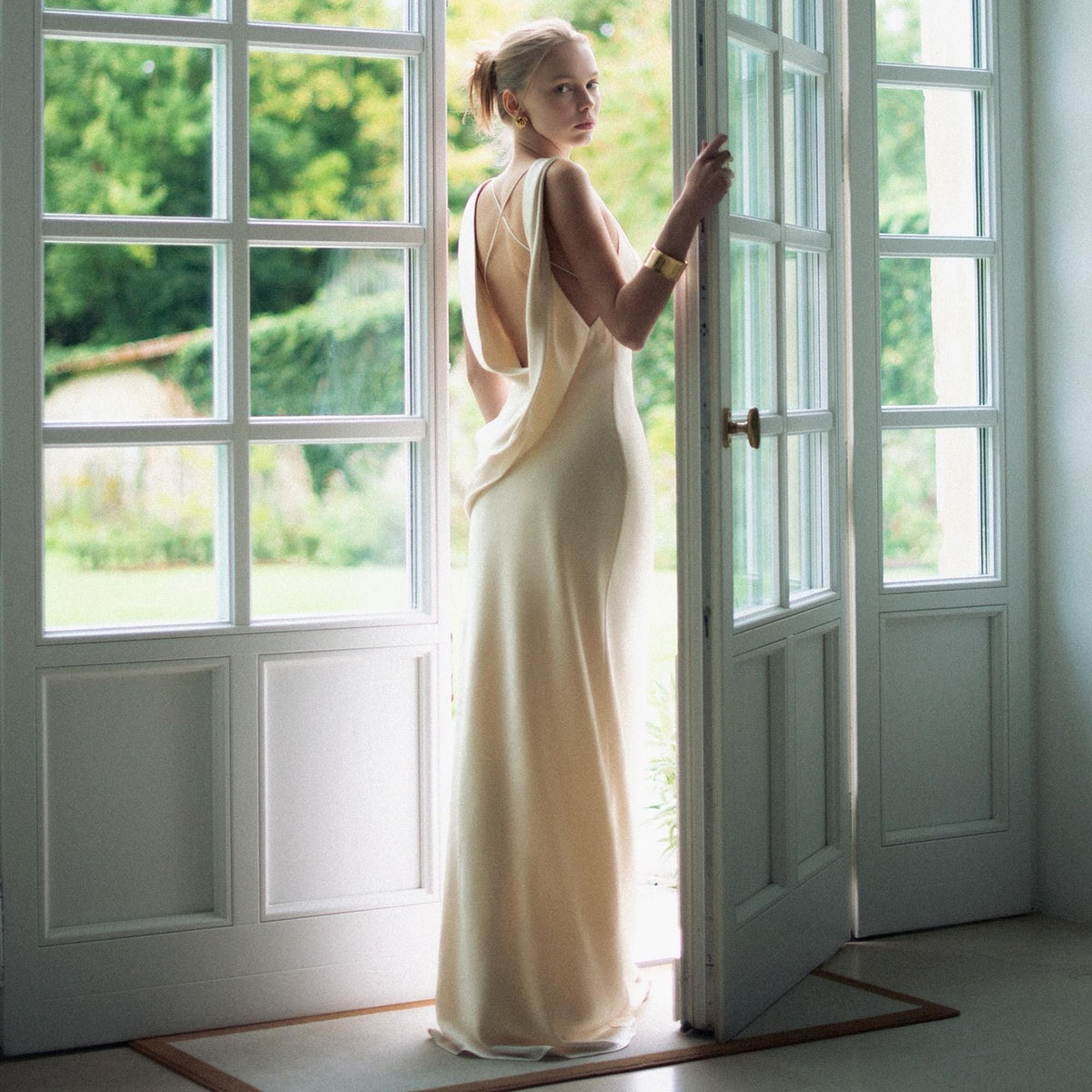 May include: A floor-length, sleeveless cream-colored gown featuring a deep cowl back design with thin, crisscrossing straps. The dress has a fluid, column-like silhouette that falls straight to the floor, made of a smooth, solid-colored fabric.
