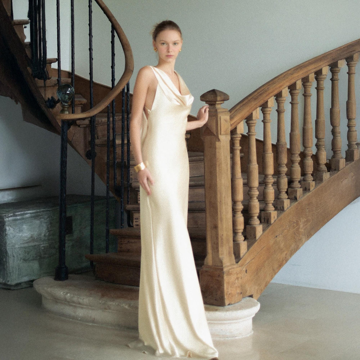 May include: A floor-length, sleeveless cream-colored satin gown featuring a draped cowl neckline and a fluid, bias-cut silhouette that falls straight to the floor. The fabric has a subtle sheen, and the dress is worn with a gold-toned cuff bracelet.