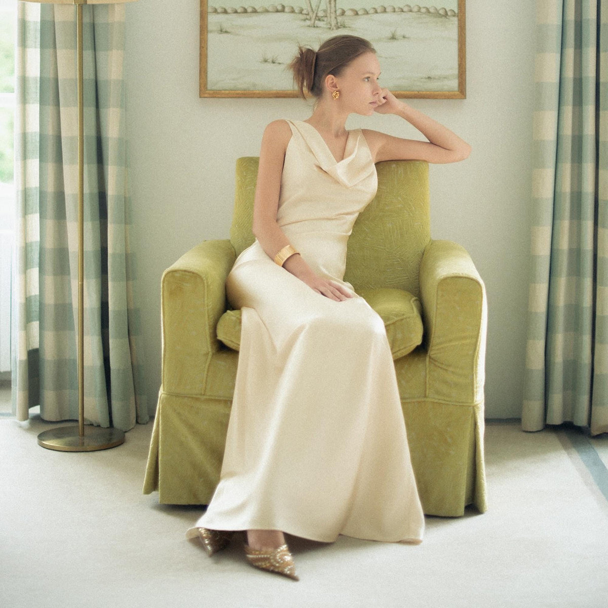 May include: A floor-length, sleeveless cream-colored gown featuring a draped cowl neckline and a fluid, bias-cut silhouette that falls straight to the floor. The dress is made of a smooth, satin-like fabric with a subtle sheen and a gold cuff bracelet.