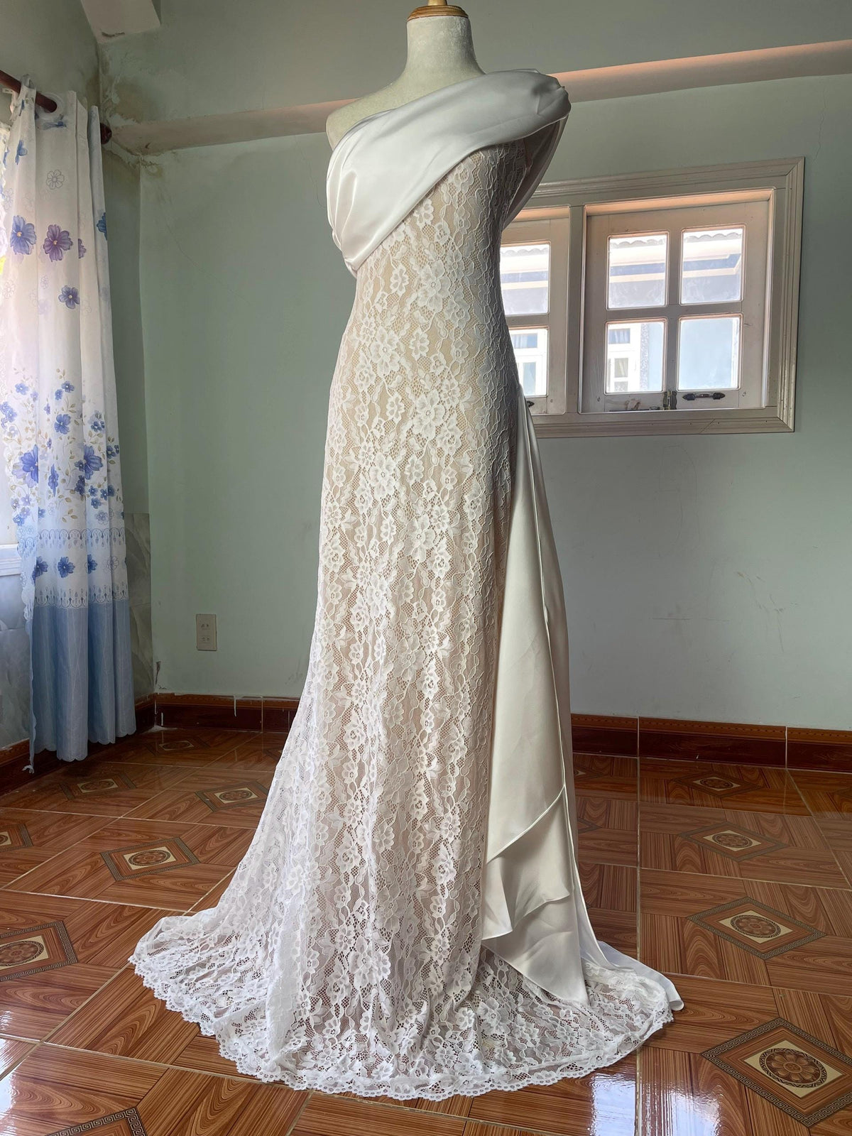 May include: A floor-length gown featuring a white lace overlay atop a beige base. The dress has an asymmetrical one-shoulder neckline with a smooth white satin drape across the chest and a matching satin panel cascading down the side.
