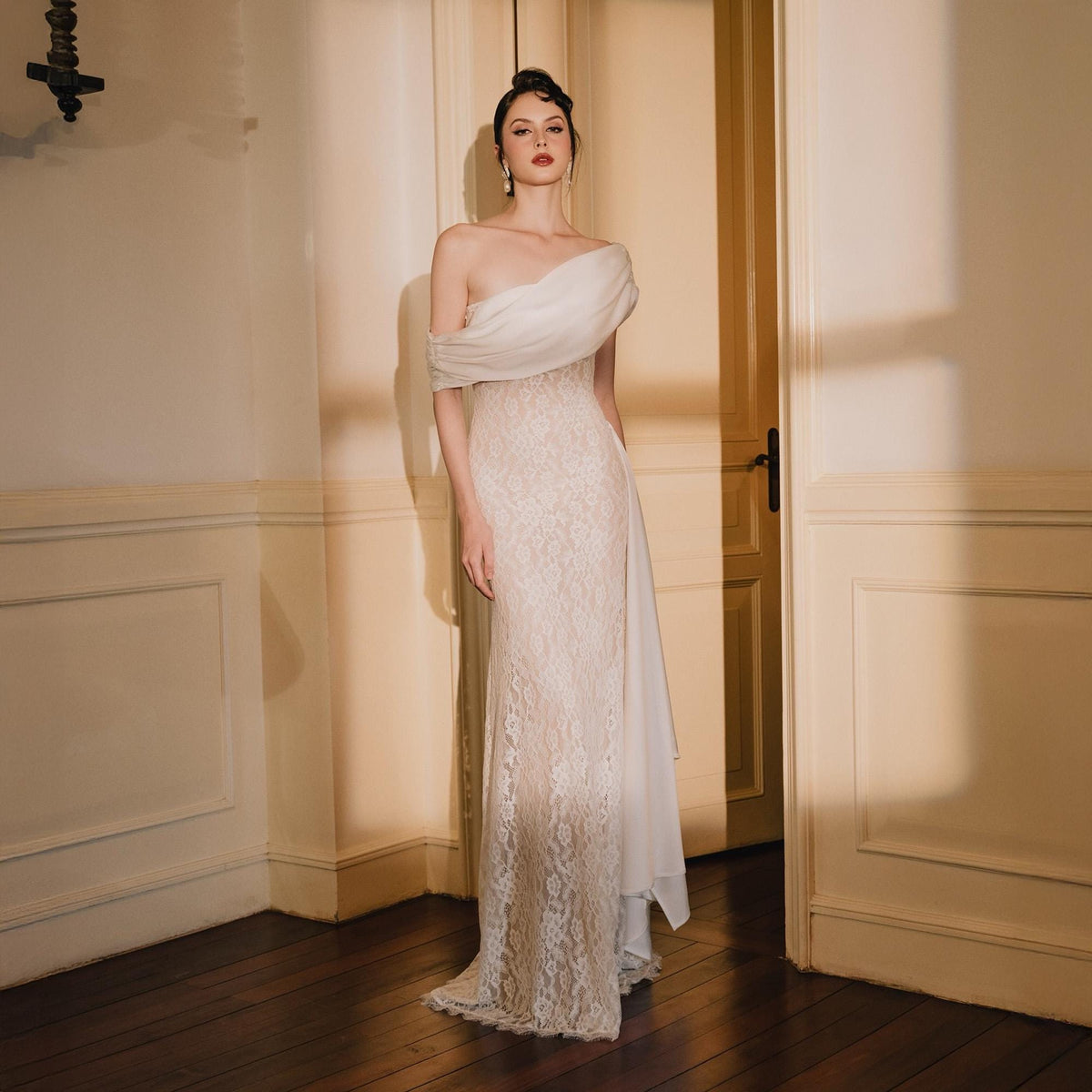 May include: A floor-length gown featuring a white, draped off-the-shoulder bodice and a fitted, cream-colored lace skirt. The garment has a straight silhouette with a sheer, flowing fabric panel extending from the side, creating a layered effect.