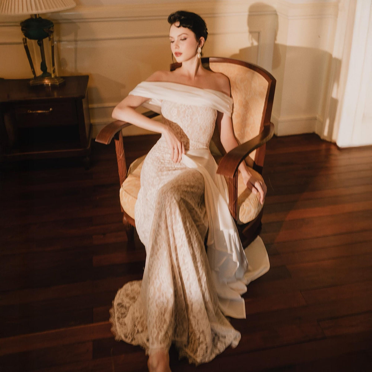 May include: An off-the-shoulder floor-length gown featuring a cream-colored lace overlay with a fitted bodice and a smooth white fabric drape across the neckline and shoulders, extending into a flowing skirt that pools on the floor.