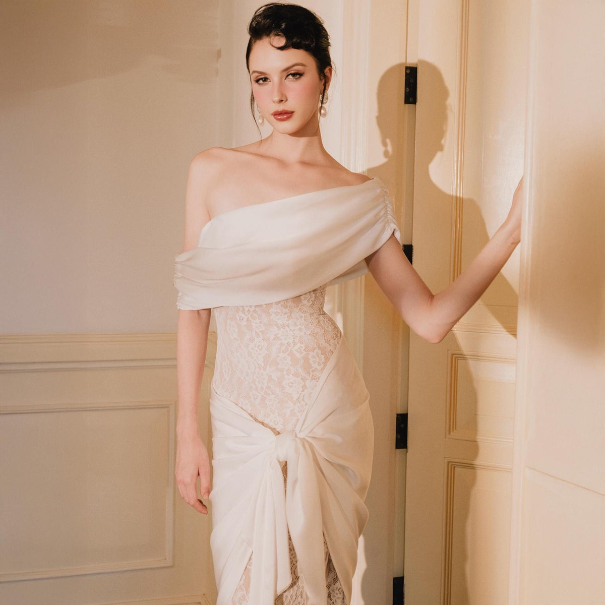 May include: A white off-the-shoulder dress featuring a lace bodice and a draped, knotted fabric overlay. The garment has a smooth, flowing neckline and a fitted lace midsection that transitions into a gathered, tied skirt detail at the front.