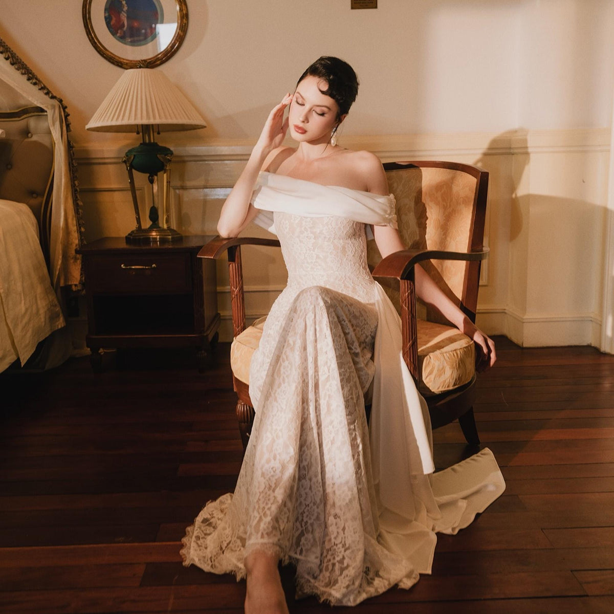 May include: A floor-length off-the-shoulder gown featuring a white lace overlay with a fitted bodice and a flowing skirt. The garment includes a sheer white fabric draped across the shoulders and upper arms, creating a soft, elegant silhouette.