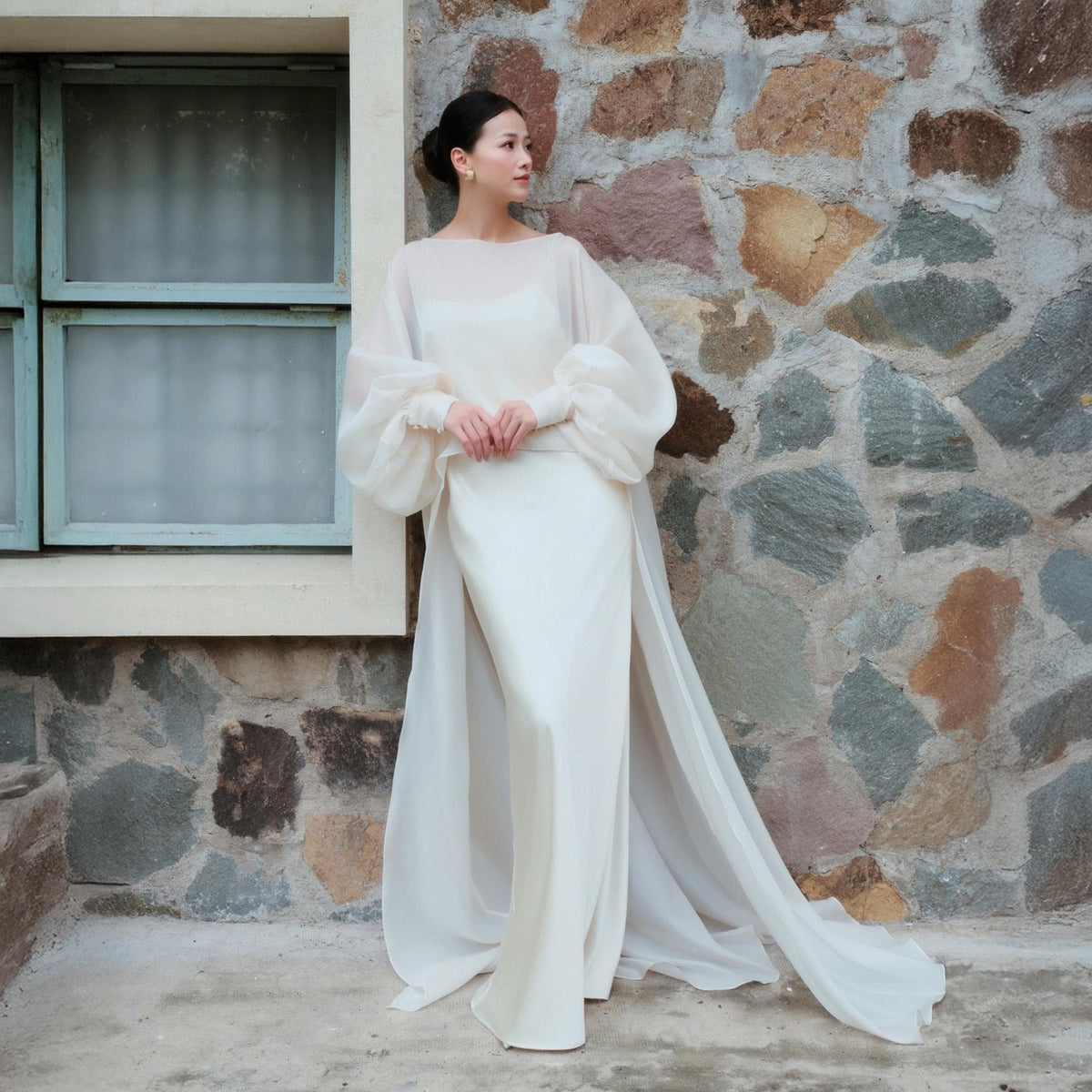 May include: A white bridal gown with a sheath silhouette and a sheer, flowing overcoat. The dress features long, sheer sleeves with gathered cuffs. The model is standing next to a stone wall and a window with blue trim.