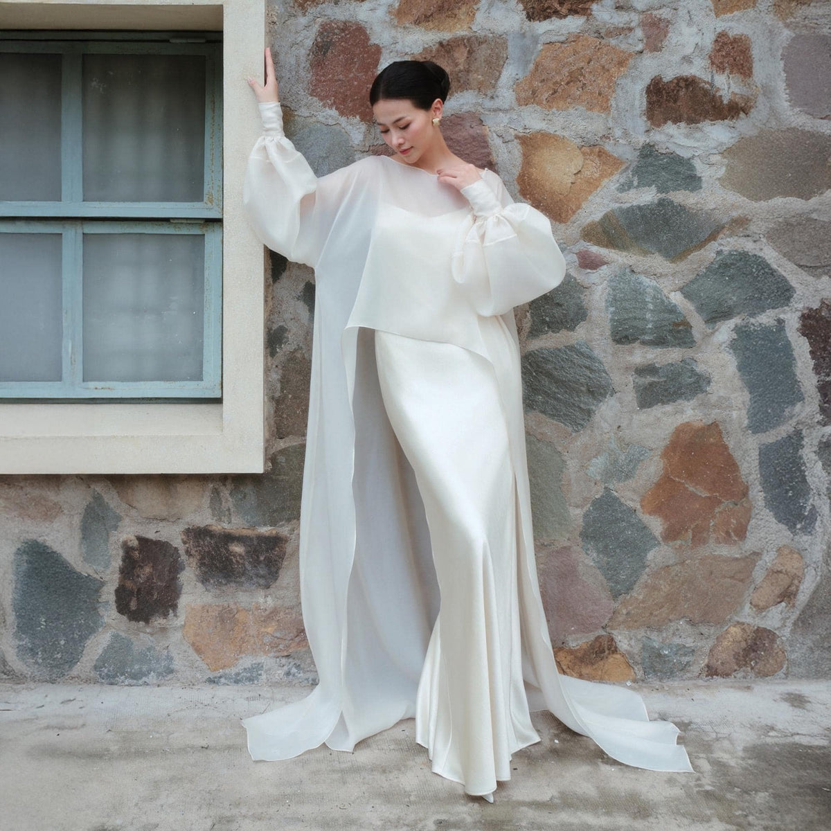 May include: A cream-colored bridal gown with a flowing, floor-length skirt and a sheer, long-sleeved overlay. The dress features a high neckline and a simple, elegant design. The model is standing next to a stone wall and a window.