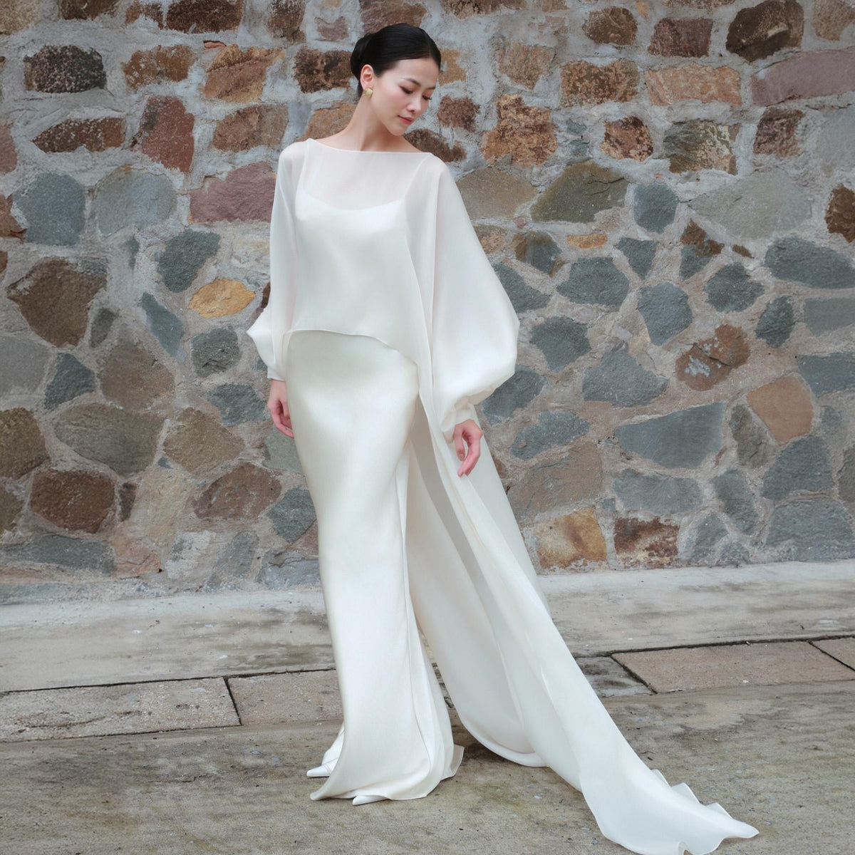 May include: A white wedding dress with a long train. The dress has a fitted silhouette with a high neckline and long sleeves. A sheer, flowing overcoat with voluminous sleeves adds a dramatic touch. The dress is made of a silky fabric.