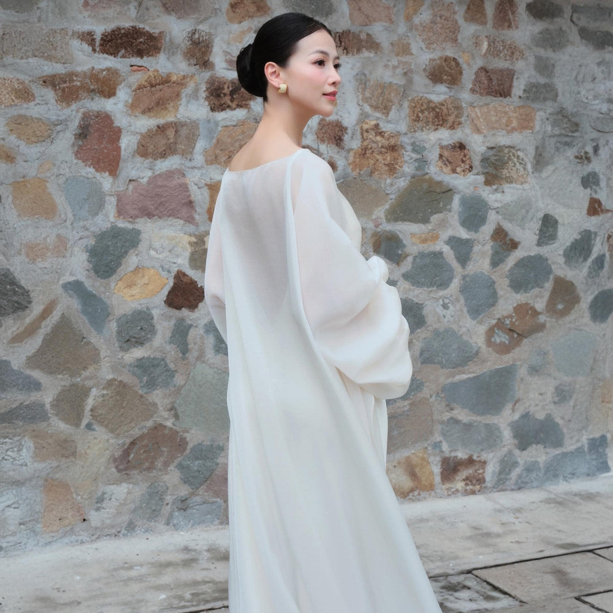 May include: A flowing, off-white dress with long, billowy sleeves. The dress has a simple neckline and a floor-length skirt. The fabric appears to be lightweight and sheer. The woman is wearing gold earrings and has her hair up in a bun.